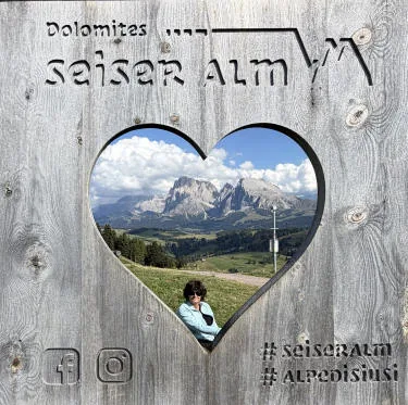 Una cornice di legno con un’apertura a forma di cuore: dentro si vedono le Dolomiti e Anna sul prato.